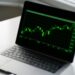 A black laptop displaying a green line graph with various peaks and troughs, likely representing financial or stock market data, sits on a white table near a window with sunlight streaming through. The chart appears to track the performance of top coins in the market. | eCryptoBit