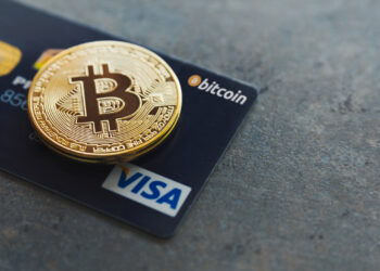 A golden Bitcoin coin rests on a dark Visa credit card, subtly hinting at the seamless integration of traditional and digital currencies. The prominent Bitcoin logo on the coin pairs with the Visa logo on the bottom right corner of the card, which also features contactless payment symbols—a subtle guide on how to buy Bitcoin. | eCryptoBit
