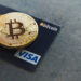 A golden Bitcoin coin rests on a dark Visa credit card, subtly hinting at the seamless integration of traditional and digital currencies. The prominent Bitcoin logo on the coin pairs with the Visa logo on the bottom right corner of the card, which also features contactless payment symbols—a subtle guide on how to buy Bitcoin. | eCryptoBit