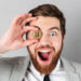 A bearded man in a light gray suit and polka dot shirt holds a gold Bitcoin close to his right eye with an excited expression, as if he's discovered the top crypto to buy. The gray background accentuates his surprise. | eCryptoBit