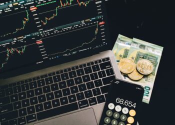 A laptop displaying stock charts, a smartphone with a calculator app showing 68,654, two cryptocurrency coins, and euro banknotes are arranged on a black surface. | eCryptoBit