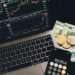 A laptop displaying stock charts, a smartphone with a calculator app showing 68,654, two cryptocurrency coins, and euro banknotes are arranged on a black surface. | eCryptoBit